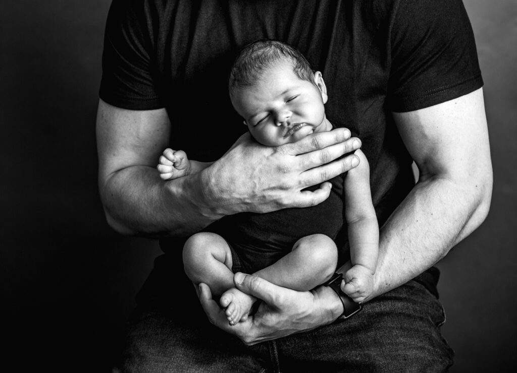 Black and white image of newborn baby in father's arms, safely posed by supporting baby's head and body.