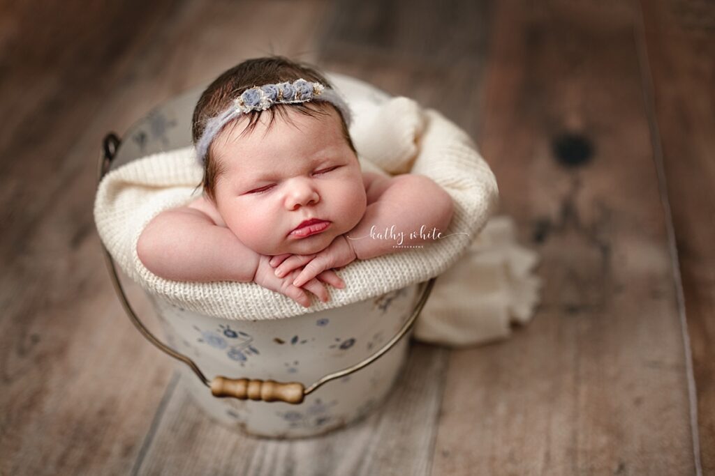 A sleeping newborn baby girl wearing a blue floral headband posed inside a floral bucket Kathy White Photography's New Market studio.