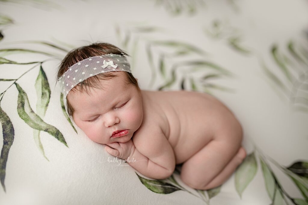 A sleeping newborn baby girl wearing a headband laying on a backdrop with greenery in Kathy White's New Market studio.