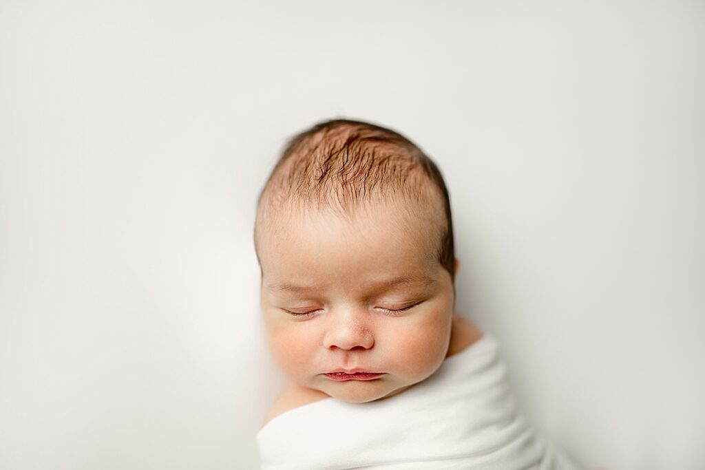 Sleeping newborn baby swaddled in white on white background.