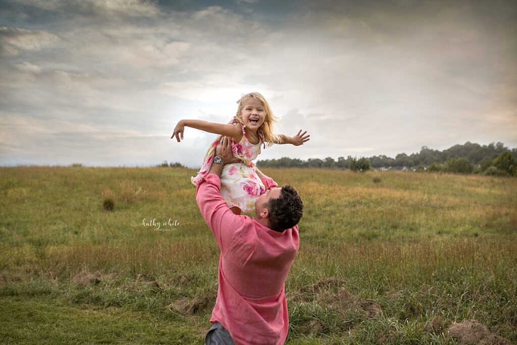 Young girl, smiling and laughing, while being tossed into the air by her dad in a field.