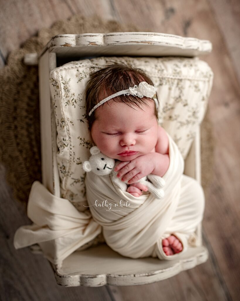 Newborn girl holding a stuffed bear, sleeping in a small crib and swaddled in a neutral fabric with toes peeking out.
