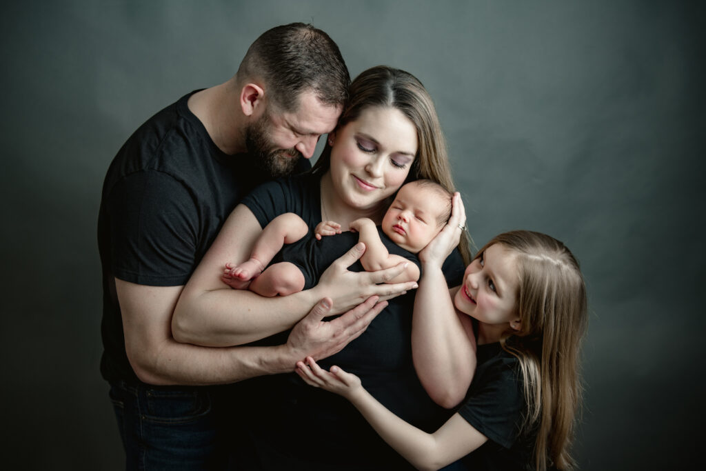 Family of four posing in a photo studio with a gray background, holding a newborn baby.